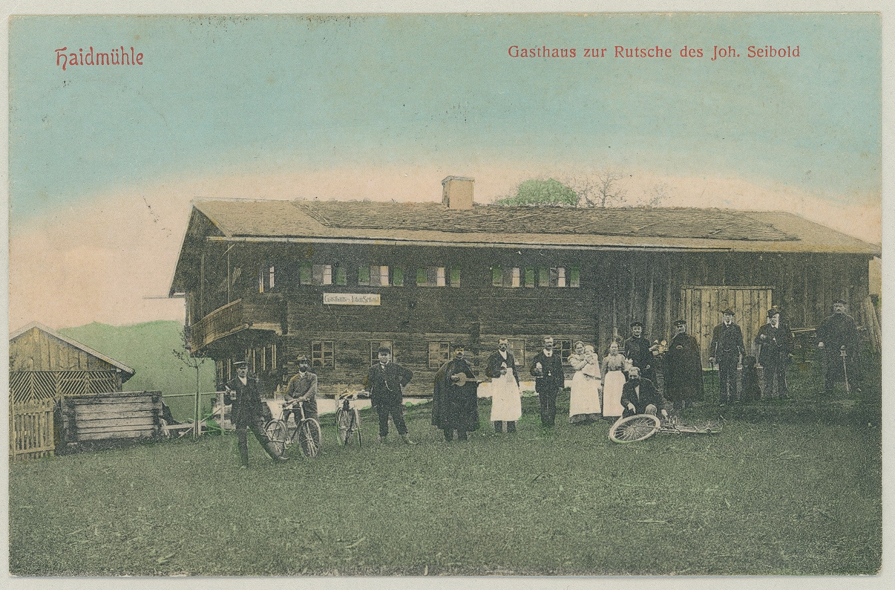 Ansicht von der Haidmühle im Landkreis Freyung-Grafenau, Gasthaus zur Rutsche: Eine Gruppe von Personen, teilweise mit Fahrrädern, posiert vor dem Gasthaus (Inhaber: Joh. Seibold). Postkarte/Fotochromolithografie, 1915